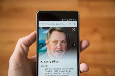 London, december 31, 2017: Larry Ellison on forbes website, mobile phone version. Hand holding smartphone with wooden background clipart