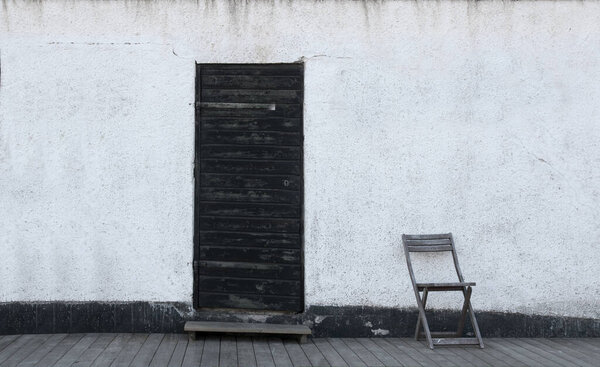 Sparse image with white wall, black vintage wooden door with peeling paint and simple wooden chair
