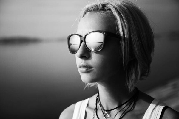 Stylish woman with short hair standing in sunglasses near river in city at summer