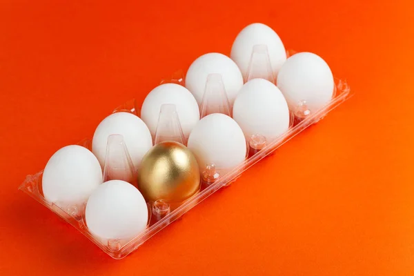 Witte kippeneieren in een pakket met een gouden ei, op een oranje achtergrond. Stockfoto
