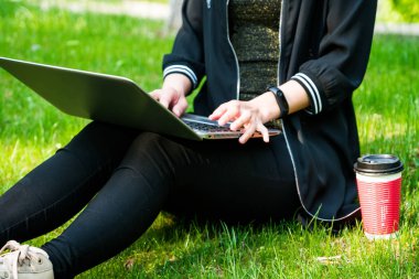 Kırmızı bir fincan kahve ile dizüstü bilgisayar kullanırken çimenlerin üzerinde oturan bir kadın