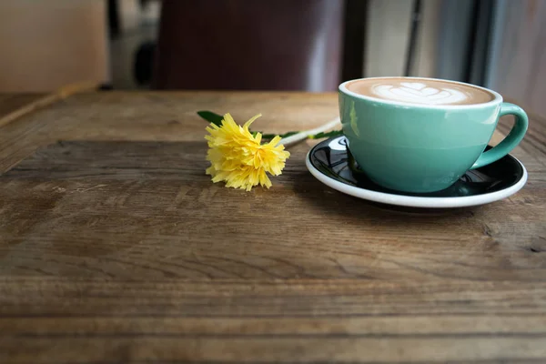 Café mocha quente ou capuchino no copo verde com padrão de coração e flor amarela na mesa de madeira — Fotografia de Stock