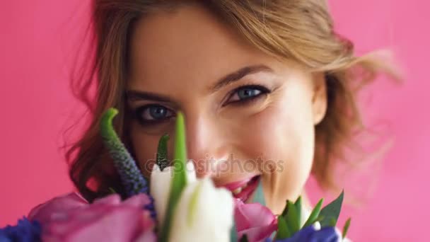 Bonita rubia joven con ojos azules y un cabello rubio largo y saludable oliendo flores sobre un fondo rosa en jeans — Vídeos de Stock