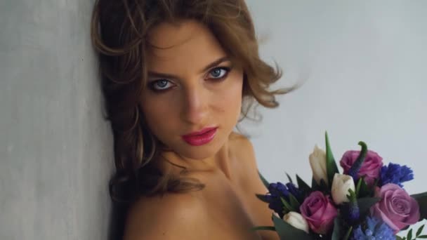 Joven hermosa mujer con sexy cuerpo delgado y vientre desnudo en un sujetador erótico de pie sosteniendo un ramo de flores sobre un fondo gris — Vídeo de stock