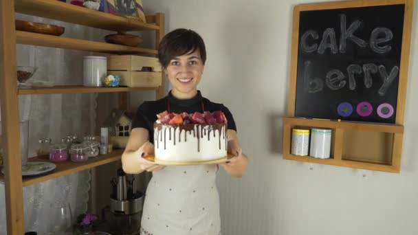 Молодая красивая женщина в фартуке делает торт со сливками и ягодами на кухне — стоковое видео