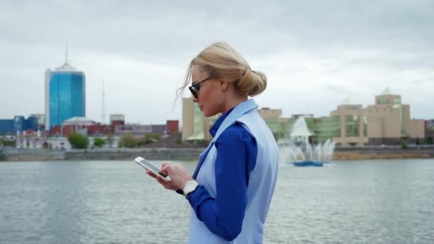 Fiatal szőke vonzó nő séta a parkban, a folyó és beszéd a mobiltelefonon. Kék póló, öltöny. — Stock videók