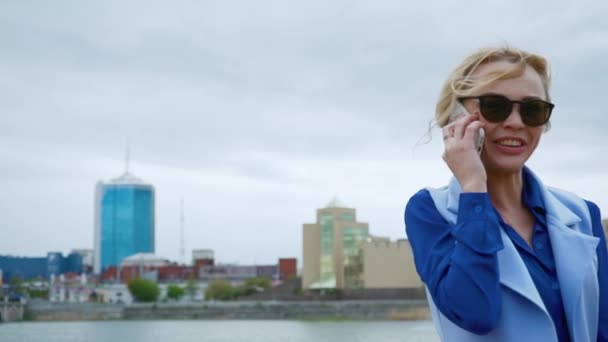 Ung blond attraktiv kvinna promenader i parken vid floden och talar på mobiltelefonen. Blå skjorta, kostym. — Stockvideo