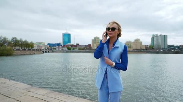 Joven rubia atractiva mujer caminando en el parque junto al río y hablando por teléfono móvil. Camisa azul, traje de negocios . — Vídeos de Stock