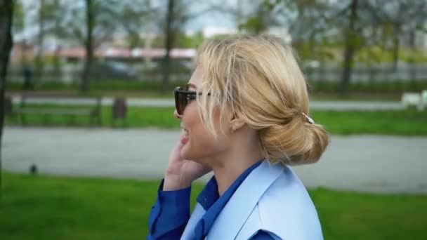 Ung blond attraktiv kvinna promenader i parken vid floden och talar på mobiltelefonen. Blå skjorta, kostym. — Stockvideo
