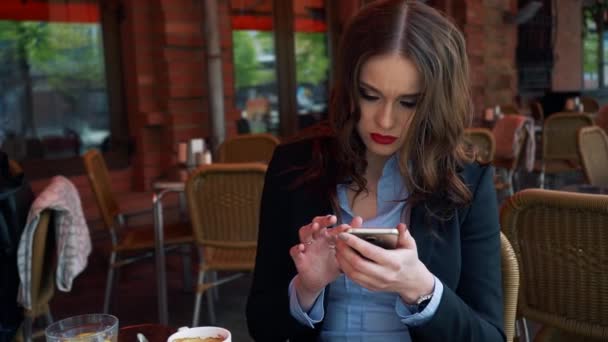 Jeune femme bien habillée avec une silhouette parfaite regardant vers son écran de téléphone intelligent pendant le petit déjeuner en terrasse du restaurant, navigation féminine sur Internet sur son téléphone à écran tactile au café — Video
