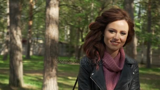 Chica en el parque, en el fondo de los árboles. Chica adolescente mira hacia abajo, luego cepilla el cabello detrás de su oreja, y mira a la cámara con una sonrisa leve, toma una bengala profunda de la lente de respiración, cámara lenta — Vídeos de Stock