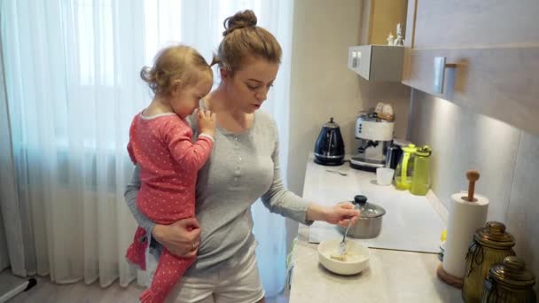 Niñera con bebé en las manos cocinar plato de comida mezcla en olla y saborearlo con cuchara . — Vídeos de Stock