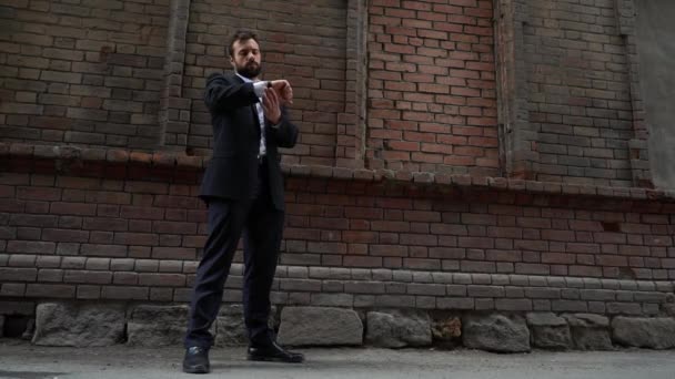 Distracted businessman pulling out his mobile phone from his pocket as he walks along an urban sidewalk alongside a brick wall with copy space — Stock Video