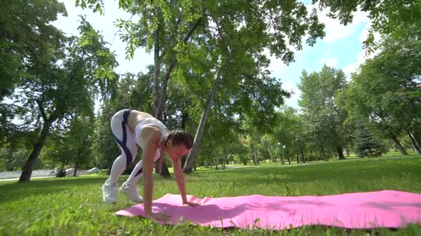 Vue latérale de la belle femme sportive slim fit à l'extérieur faire des pompes exercice et entraînement en plein air dans le parc sur un tapis de yoga — Video