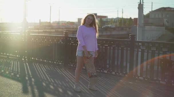Дівчина скейтбордистка, яка стоїть зі скейтбордом на заході сонця — стокове відео