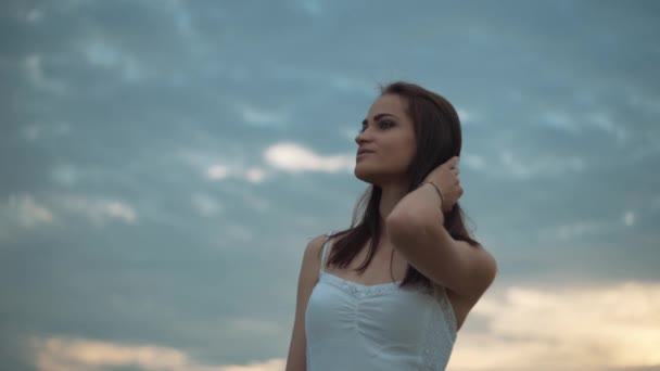 Retrato de uma jovem mulher com cabelos longos em vestido branco em pé no campo do trigo e olhando para a câmera — Vídeo de Stock