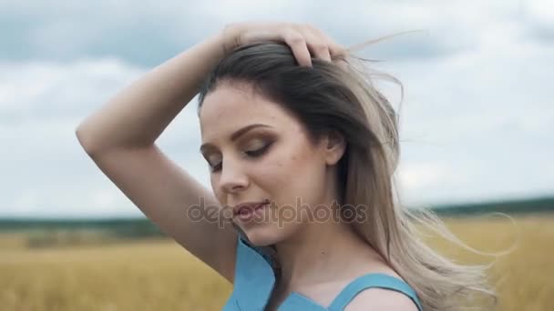 Uma jovem com um vestido azul está de pé no campo, e o vento está balançando seu cabelo — Vídeo de Stock