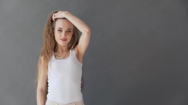 Hermosa joven con cabello rubio saludable. En una camiseta blanca sobre un fondo gris, posando sobre la cámara . — Vídeos de Stock
