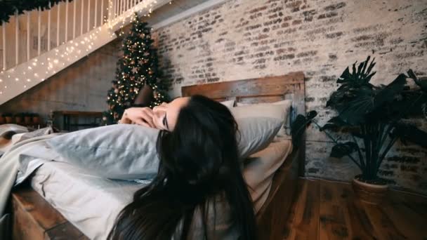 Сексуальная женщина с темными волосами в кружевном костюме лежит на кровати. На заднем плане дерево, гирлянды. Новый год, Рождество . — стоковое видео
