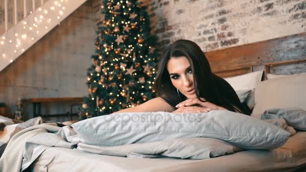 Mujer sexy con el pelo oscuro en un traje de encaje acostado en la cama. Al fondo, árbol, guirnaldas. Año Nuevo, Navidad . — Vídeo de stock