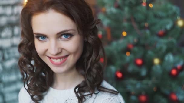 Ein schönes junges Mädchen in einem Pullover und Strümpfen sitzt neben schönen Weihnachtsbäumen und hält in den Händen ein Geschenk, Neujahr Home Interieur — Stockvideo