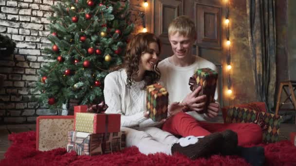 Jóvenes felices se regalan unos a otros junto a la chimenea cerca del árbol de Navidad. — Vídeos de Stock