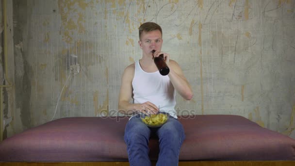 Joven bebiendo cerveza en el sofá de casa y viendo televisión — Vídeo de stock
