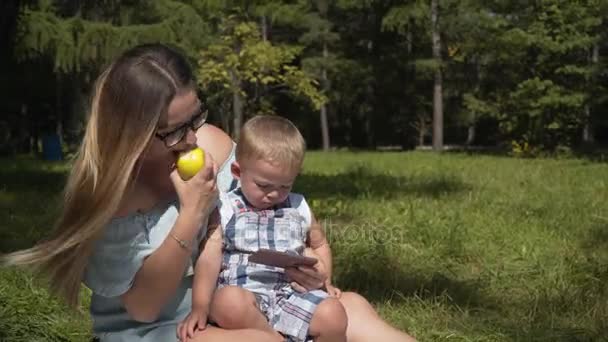 Mãe e criança em um piquenique no parque olhando smartphone e comer uma maçã . — Vídeo de Stock