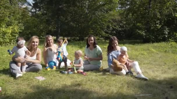 Група щасливих дітей, які грають на відкритому повітрі в літньому парку. Матері доглядають за своїми дітьми, сидячи на траві . — стокове відео