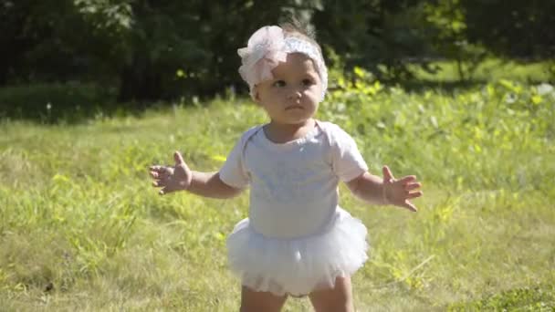 Schattig een jaar oud meisje maakt haar eerste stappen buiten. Slow Motion Video Beelden van de eerste stappen van het kind. Zonnige dag en groen gras — Stockvideo