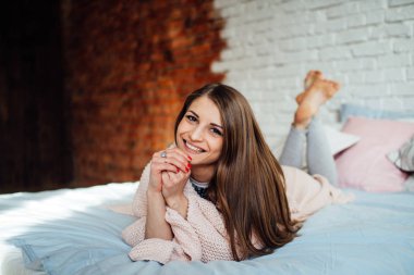 Güzel bir genç esmer hafif gömlekli modern yatak odasında yatakta yalan süre gülüyor. Sevimli kız evde kışın dinlenme.