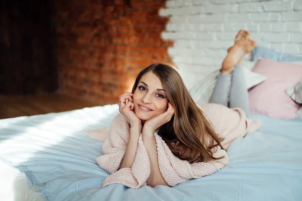 Güzel bir genç esmer hafif gömlekli modern yatak odasında yatakta yalan süre gülüyor. Sevimli kız evde kışın dinlenme. — Stok fotoğraf