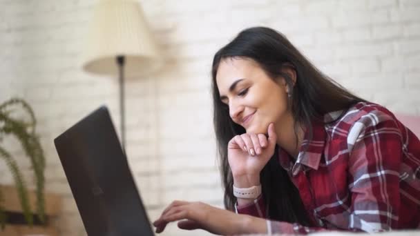 Jonge vrouw achter laptop thuis werken. Gekleed in een plaid shirt, tegen een bakstenen muur achtergrond. — Stockvideo