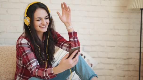 Mujer joven con auriculares, mensajes de texto en el teléfono inteligente. La chica habla por teléfono vía video. La mujer de los auriculares amarillos — Vídeos de Stock