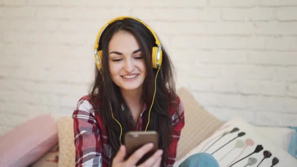 Mujer joven con auriculares, mensajes de texto en el teléfono inteligente. La chica habla por teléfono vía video. La mujer de los auriculares amarillos — Vídeos de Stock