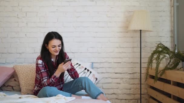 Chica joven sentada en la cama y viendo contenido en el teléfono inteligente — Vídeos de Stock