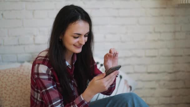 Chica joven sentada en la cama y viendo contenido en el teléfono inteligente — Vídeos de Stock