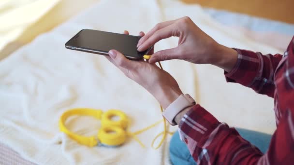 Chica sentada en la cama conecta los auriculares al teléfono . — Vídeo de stock