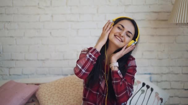 The girl listens to music with headphones and sitting on the bed dancing — Stock Video