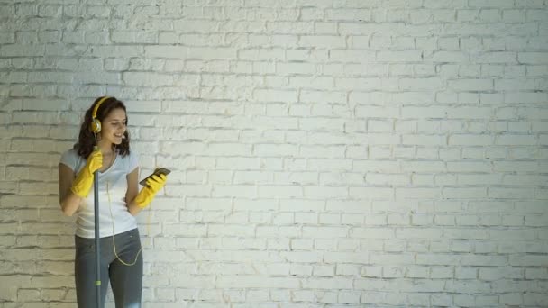 Happy woman in headphones singing, pretending to MOP the microphone, amid the white brick wall — Stock Video