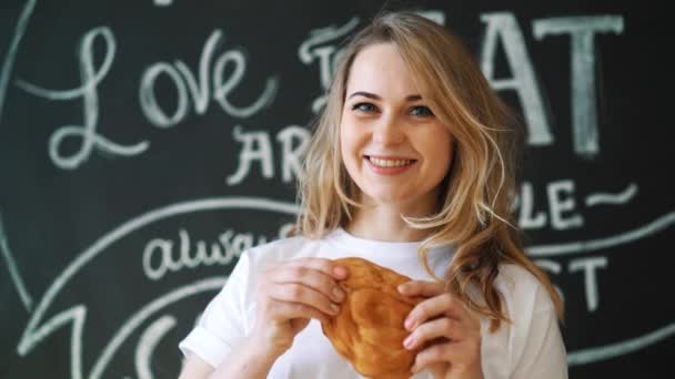 Giovane bella ragazza in cucina si siede a un tavolo sta tenendo una pagnotta guardando la fotocamera e sorridendo. Ritratto di una bella ragazza nella sala da pranzo a colazione . — Video Stock