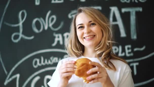 Joven chica hermosa en la cocina se sienta en una mesa está sosteniendo una barra de pan mirando a la cámara y sonriendo. Retrato de una chica atractiva en el comedor en el desayuno . — Vídeos de Stock