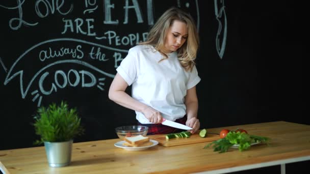 Belle jeune femme coupe concombre vert avec couteau de cuisine sur des morceaux ronds sur planche à découper en bois et prépare une salade fraîche saine, à côté il y a des feuilles de laitue verte et de tomate avec concombre sur — Video