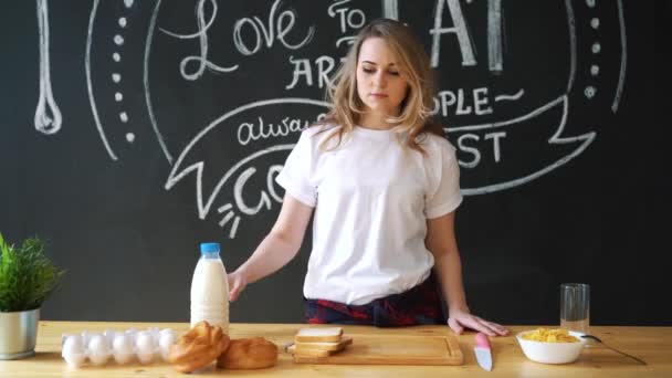 Una bella donna vestita in pigiama, una t-shirt bianca, farà colazione a casa in cucina. Latte, panino, fiocchi — Video Stock