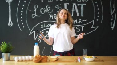 Genç ve çekici bir kadın sabah mutfakta omlet pişiriyor, gülümsüyor, mutlu pozitif ev hanımı, sağlıklı yaşam tarzı, kulaklıkla müzik dinliyor, gülüyor, eğleniyor, dans ediyor, şarkı söylüyor.