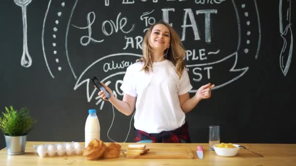 Jovem atraente mulher cozinhar ovos mexidos na cozinha de manhã, sorrindo, feliz dona de casa positiva, estilo de vida saudável, ouvir música em fones de ouvido, rindo, se divertindo, dançando, cantando — Vídeo de Stock