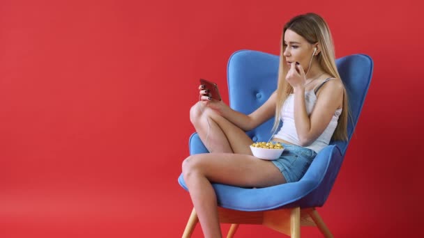 La chica se sienta en una silla, viendo una película por teléfono y comiendo palomitas de maíz — Vídeos de Stock