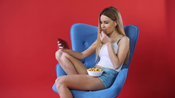 La chica se sienta en una silla, viendo una película por teléfono y comiendo palomitas de maíz — Vídeos de Stock