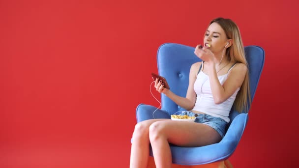 La chica se sienta en una silla, viendo una película por teléfono y comiendo palomitas de maíz — Vídeos de Stock