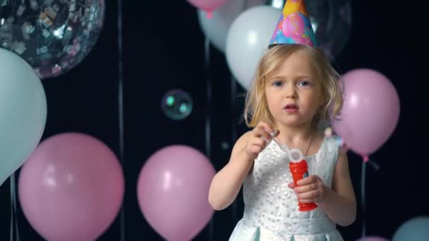 Маленькая девочка надувает пузыри на фестивале, на фоне воздушных шаров и конфетти . — стоковое видео